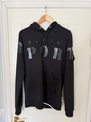 Svart hoodie från Plein Sport - Snygg svart hoodie från Plein Sport med stora, glansiga bokstäver på bröstet och ärmarna. Klassisk känguruficka framtill och justerbar huva med snören. Tillverkad i mjukt material som passar perfekt till en streetwear-look.