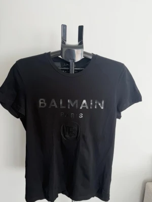 Svart Balmain Paris t-shirt - Snygg svart t-shirt från Balmain Paris med glansigt tryck framtill. Klassisk rund halsringning och korta ärmar. Tillverkad i mjuk bomull för en bekväm känsla. Perfekt för dig som gillar exklusiva streetwear-plagg.