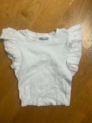 Vit topp med volangärmar från Zara - Säljer en vit ribbad topp från Zara med korta volangärmar och rund halsringning. Materialet är mjukt och stretchigt, perfekt för en trendig och enkel look. Passar till både jeans och kjol för en fräsch stil.