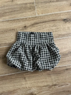 Svartvit rutig kortkjol Zara - Trendig svartvit rutig kortkjol från Zara med hög midja och volangig form. Kjolen har ett klassiskt hundtandsmönster och är tillverkad i ett tjockare tyg som ger struktur. Perfekt för dig som vill sticka ut med en cool och chic look.