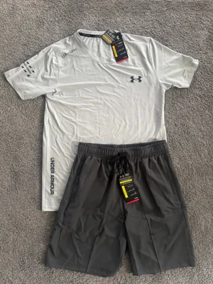 Under Armour träningsset grå/svart - Snyggt träningsset från Under Armour med en ljusgrå t-shirt och mörkgrå shorts. T-shirten har tryckta loggor och textdetaljer, medan shortsen har elastisk midja med dragsko och färgglada loggor. Perfekt för gymmet eller löpning. Materialet är lätt och andas.