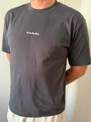 Mörkgrå Acne Studios t-shirt - Snygg mörkgrå t-shirt från Acne Studios med enkel vit logga på bröstet. Klassisk passform med rund hals och korta ärmar. Tillverkad i mjuk bomull som känns skön mot huden. Perfekt för dig som gillar minimalistisk och stilren design.