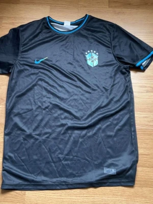 Svart Brasilien Nike fotbollströja - Snygg svart fotbollströja från Nike med Brasilien-emblem och fem stjärnor på bröstet. Tröjan har blå detaljer vid krage och ärmslut samt Nike-logga på framsidan. Tillverkad i lätt och ventilerande material, perfekt för träning eller match.