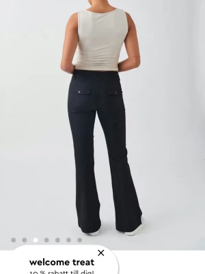 Svarta bootcut-byxor med fickor - Snygga svarta bootcut-byxor med två bakfickor med lock och knapp. Byxorna har hällor för skärp och är tillverkade i ett stretchigt material som ger en bekväm passform. Perfekta för en trendig och avslappnad stil.