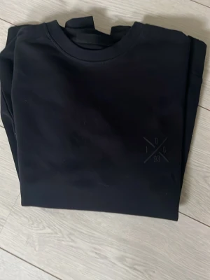 Svart sweatshirt från Indicode - En stilren svart sweatshirt från Indicode med rund halsringning och diskret broderad logga på bröstet. Tillverkad i mjukt material som passar perfekt till jeans eller joggers. Enkel och clean design för en avslappnad look.