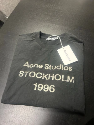 Svart Acne Studios t-shirt Stockholm - Svart t-shirt från Acne Studios med tryck i vitt framtill där det står 'Acne Studios STOCKHOLM 1996'. Klassisk rund halsringning och korta ärmar. Perfekt för dig som gillar stilrena och ikoniska plagg.