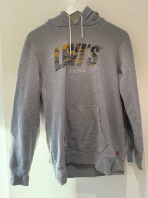 Grå Levi's hoodie med tryck - Säljer en grå hoodie från Levi's med coolt kamouflagemönstrat tryck i gult och grått på bröstet där det står 'Levi's California'. Hoodien har huva med vita snören och en stor magficka. Perfekt för dig som gillar streetwear och avslappnad stil.