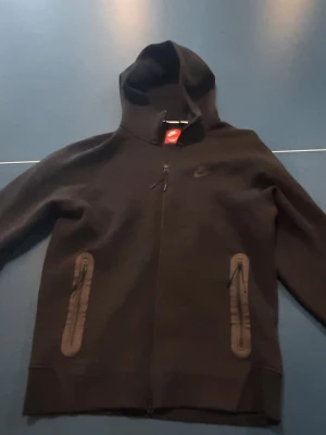 Svart Nike hoodie med dragkedja - Svart hoodie från Nike med hel dragkedja framtill och två fickor med dragkedja. Klassisk huva och diskret Nike-logga på bröstet. Tillverkad i mjukt material som känns skönt mot huden. Perfekt för en sportig och avslappnad stil.