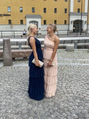 Ljusrosa långklänning (balklänning) med volanger - (Den högra klänningen) Elegant ljusrosa långklänning med flera lager volanger och breda axelband. Klänningen har en figurnära passform upptill och faller snyggt längs kroppen. Perfekt för speciella tillfällen. Jättefin till bal! Storlek 38