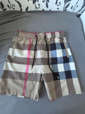 Burberry shorts Rutig shorts med snörning - Snygga shorts med klassiskt rutigt mönster i beige, brunt, svart, vitt och en rosa detalj. Shortsen har elastisk midja med svart snörning och sidofickor. Perfekta för sommaren och ger en trendig look.