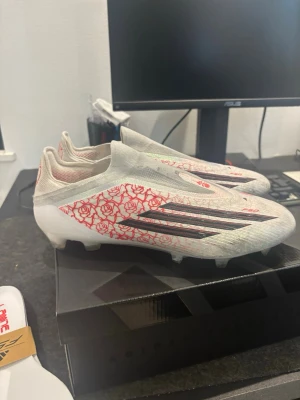F50 Lamine yamal  - Snygga Adidas X Speedflow fotbollsskor i vitt med röda rosdetaljer och svarta stripes. Skorna har låg vikt, strumpliknande passform och är tillverkade i syntetmaterial för bästa känsla på planen. Perfekta för dig som vill sticka ut på matchen.