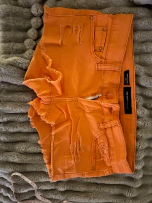 Orange slitna shorts från miss sixty  - Säljer ett par orangea shorts från miss sixty med slitna detaljer och råa kanter. Shortsen har fem fickor, dragkedja och knapp framtill. Perfekta för sommaren och ger en avslappnad vibe.