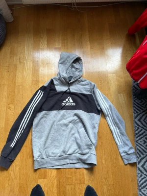 Grå och svart hoodie från Adidas - Snygg hoodie från Adidas i grått och svart med klassiska vita ränder längs ärmarna och stor logga på bröstet. Huva med dragsko och magficka framtill. Perfekt för en sportig och avslappnad stil.