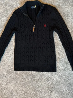 Ralph Lauren Half Zip - Svart kabelstickad tröja från Polo Ralph Lauren med halv dragkedja och ribbad krage. Tröjan har ett rött broderat logotyp på bröstet och ribbade muddar. Storlek M