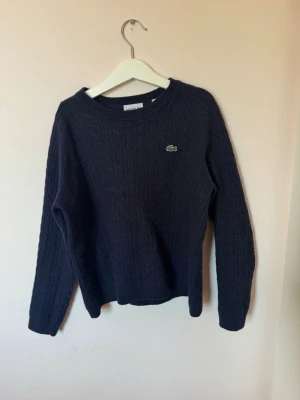 Mörkblå kabelstickad tröja Lacoste - Mörkblå kabelstickad tröja från Lacoste med rund halsringning och den klassiska gröna krokodilloggan på bröstet. Tröjan har långa ärmar och ribbade muddar vid ärmslut och nederkant. Perfekt för dig som gillar stilrena och tidlösa plagg. Storlek 152 men är liten i storlek, passar 140 