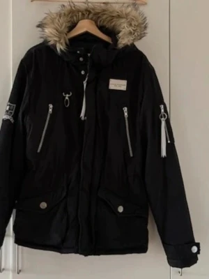 Svart parkas med fuskpäls från Svea - Snygg svart parkas från Svea med avtagbar huva och fuskpäls. Jackan har dragkedjor i silver, flera fickor och justerbar midja. Perfekt för kalla dagar med sin varma och stilrena look. Svea-logga på bröstet och ärmen ger extra streetkänsla.