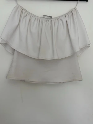 Vit offshoulder topp med volang från Zara - Säljer en vit axelbandslös topp med bred volangkant och offshoulder-design. Toppen har en luftig och flowy passform, perfekt för varma dagar. Materialet känns mjukt och lätt mot huden.