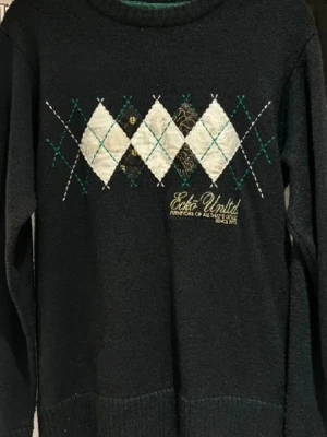 Svart argyletröja från Ecko Unltd - Snygg svart stickad tröja från Ecko Unltd med klassiskt argylemönster i vitt, beige och grönt framtill. Broderad logga i gult nedtill. Tröjan har rund halsringning och är perfekt för dig som gillar streetwear med en twist.