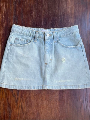 Ljusblå jeanskjol  - Trendig ljusblå jeanskjol i klassisk femficksmodell med bälteshällor och knappstängning framtill. Kjolen har en kort passform och är tillverkad i bomull. Snygga slitningar framtill ger en cool och avslappnad vibe.