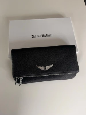 Svart kuvertväska Zadig & Voltaire - Snygg svart kuvertväska från Zadig & Voltaire i strukturerat läder. Väskan har silverfärgade detaljer, dubbla dragkedjor och en ikonisk vinglogga framtill. Perfekt för dig som gillar stilrena accessoarer med en edgy touch.