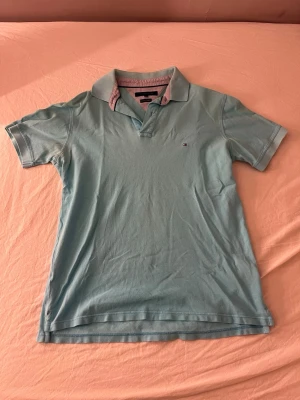 Ljusblå pikétröja Tommy Hilfiger - Säljer en ljusblå pikétröja från Tommy Hilfiger med klassisk krage och liten logga på bröstet. Tröjan är i mjuk bomull och har korta ärmar samt kontrastfärgad insida på kragen. Perfekt för en avslappnad men snygg stil.