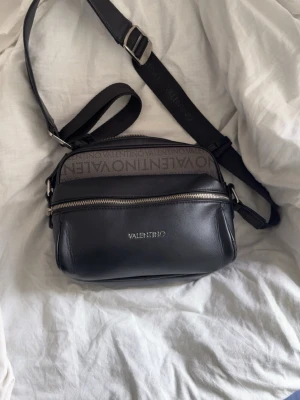 Svart axelväska från Valentino - Snygg svart axelväska från Valentino med justerbar axelrem och silverfärgade dragkedjor. Väskan har logotypen tryckt på framsidan och ett band med Valentino-tryck upptill. Tillverkad i slätt konstläder med stilren, rektangulär form.