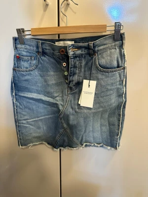 Blå jeanskjol från Scotch & Soda - Snygg blå jeanskjol från Scotch & Soda med råa kanter och klassiska fem fickor. Kjolen har knappar framtill och en liten röd detalj vid fickan. Perfekt för en avslappnad och trendig look.