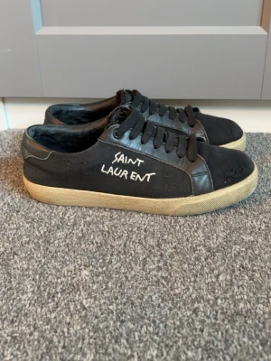 Svarta Saint Laurent sneakers - Svarta sneakers från Saint Laurent med vit logga på sidan. Skorna har lågt skaft, svarta skosnören och beige sula. Ovandelen är i en mix av läder och textil för en cool och avslappnad look. Vänstra skon har ett litet hål mellan sulan och själva skon, tveka inte på att höra av er hålet är även lätt att laga med tillexempel lim. Priset är inte hugget i sten!