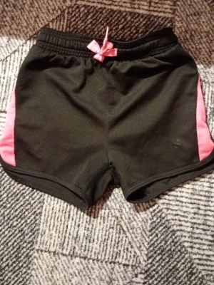 Svarta shorts med rosa detaljer - Snygga svarta shorts med rosa sidor och en liten rosa rosett i midjan. Shortsen har elastisk midja och är tillverkade i mjukt bomullsmaterial, perfekta för träning eller chill. Sportig och bekväm passform.