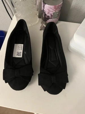 Svarta ballerinaskor med rosett - Stilrena svarta ballerinaskor i mockaimitation med rund tå och dekorativ rosett framtill. Skorna har låg sula med diskret mönster undertill för bättre grepp. Perfekta för en trendig och enkel look.
