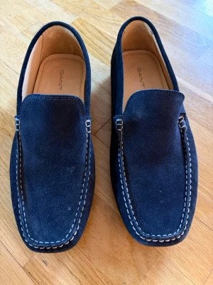 Mörkblå mocka loafers från Gant - Säljer ett par stilrena mörkblå loafers från Gant i mjuk mocka. Skorna har kontrasterande vita sömmar och klassisk slip-on design med rundad tå. Insidan är ljusbrun och ger en snygg kontrast till det mörka yttermaterialet.