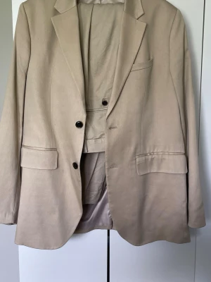Beige kavaj med klassisk passform - Stilren beige kavaj med två knappar framtill och klassiska slag. Kavajen har två fickor med lock och en bröstficka. Byxorna är relaxed fit vilket ger en askön look. 