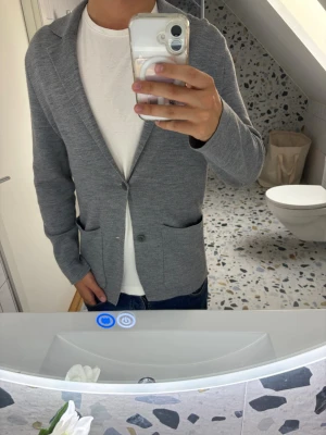 Grå stickad kavaj med två fickor från COS - Snygg grå stickad kavaj med två framfickor och enkelknäppning. Kavajen har en avslappnad passform och klassisk krage, perfekt att slänga över en t-shirt för en stilren look. Materialet är mjukt och bekvämt, vilket gör den enkel att bära hela dagen.