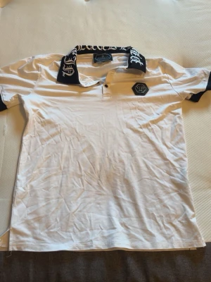 Vit Philipp Plein t-shirt med tryck - Snygg vit t-shirt från Philipp Plein med svart krage och ärmslut. Kragen har ett coolt vitt tryck och på bröstet sitter en svart logga. Klassisk passform och tillverkad i mjuk bomull, perfekt till vardagslooken.