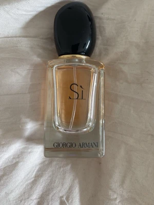 Giorgio Armani Sì parfym - Elegant parfym från Giorgio Armani, Sì, i en genomskinlig glasflaska med svart rundad kork. Flaskan har en stilren design med guldtonad vätska inuti och tydlig logga på framsidan. Perfekt för dig som gillar exklusiva dofter och snygg förpackning.