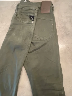 Olivgröna jeans från Replay - Snygga olivgröna jeans från Replay med klassisk femficksdesign och diskret logga på bakfickan. Jeansen har raka ben och är tillverkade i ett mjukt bomullsmaterial med lite stretch för extra komfort. Perfekta för en avslappnad och trendig look. Ny tvättade 