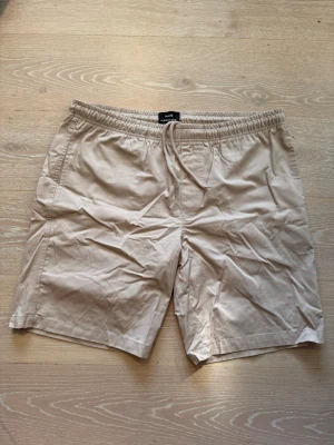 Beige shorts från Mads Nørgaard - Snygga beige shorts från Mads Nørgaard med elastisk midja och snörning. Perfekta för varma dagar, tillverkade i mjuk bomull för en skön känsla. Klassisk och enkel design som funkar till det mesta.