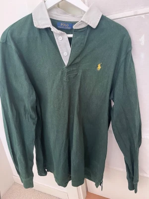 Grön henley från Polo Ralph Lauren - Klassisk grön henley från Polo Ralph Lauren med kontrasterande vit krage och knappar. Långärmad modell i bomull med den ikoniska gula logotypen broderad på bröstet. Perfekt för en avslappnad men ändå stilren look.