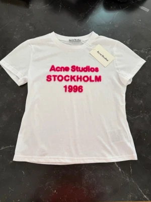 Vit Acne Studios t-shirt med rosa tryck M - Vit t-shirt från Acne Studios med rosa text 'Acne Studios STOCKHOLM 1996' på bröstet. Klassisk rund hals och korta ärmar. Tillverkad i mjuk bomull för en skön och avslappnad passform. Perfekt för dig som gillar stilrena plagg med en twist.