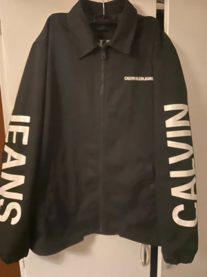 Calvin Klein Jeans svart limited jacka  - Limited svart vindjacka som jag köpte i Usa från Calvin Klein Jeans med stora vita logotryck på ryggen och ärmarna. Jackan har dragkedja framtill, klassisk krage och lång ärm. Perfekt för dig som gillar streetwear och tydliga loggor. Ordinarie pris 3400 kronor.