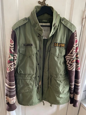 Grön parkajacka från Ralph Lauren - Unik grön parkajacka från Ralph Lauren med stickade, mönstrade ärmar i brunt, vitt, rött och grönt. Jackan har flera fickor, axelklaffar och dragkedja framtill. Märken med 'R. Lauren' och 'D.S. USRL' på bröstet ger en cool militärvibe.