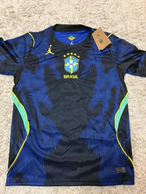 Brasilien Bortaställ - Snygg blå och svart träningströja från Nike med Brasilien CBF-logga och gula detaljer. Tröjan har korta ärmar, rund hals och mönstrad design med inslag av grönt och gult på sidorna. Perfekt för fotbollsträning eller sportiga aktiviteter.