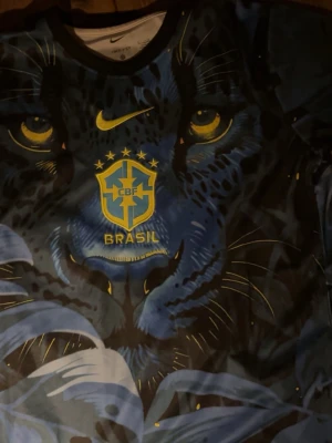 Nike Brasil t-shirt med pantermotiv - Snygg t-shirt från Nike med Brasil-logga och ett stort blått pantermotiv över hela framsidan. T-shirten har korta ärmar och rund halsringning. Färgerna är främst blå och svart med gula detaljer. Perfekt för dig som gillar unika och sportiga plagg.pris diskuteras 