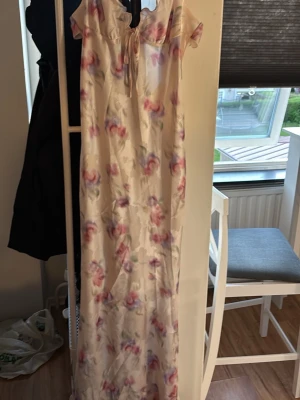 Lång blommig klänning med snörning - Romantisk långklänning i ljus bas med blommigt mönster i rosa och lila toner. Klänningen har offshoulder-ärmar och snörning framtill samt en öppen rygg med knyt. Perfekt för sommarens alla tillfällen.