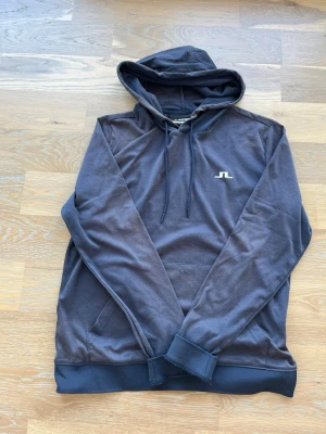 Mörkblå hoodie från J.Lindeberg - Snygg mörkblå hoodie från J.Lindeberg med klassisk huva och dragsko. Tröjan har en liten vit logga på bröstet och en känguruficka framtill. Tillverkad i mjukt material som känns skönt mot huden. Perfekt för en avslappnad och stilren look.