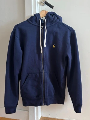 Mörkblå zip hoodie från Polo Ralph Lauren strl S - Mörkblå hoodie med dragkedja från Polo Ralph Lauren. Köptes för ca 6 månader sedan, men växtes ur snabbt. Den är i storlek S för herr, men drar mer åt XS. Klassisk design med vit dragsko i huvan,  gul broderad logga på bröstet.
