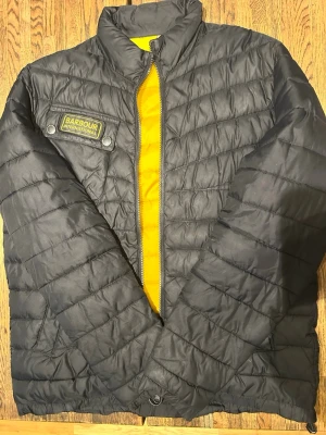 Svart pufferjacka från Barbour - Snygg svart pufferjacka från Barbour International med gul insida och patch på bröstet. Jackan har ståkrage, dragkedja och två fickor framtill. Perfekt för kalla dagar och har en modern, sportig look.