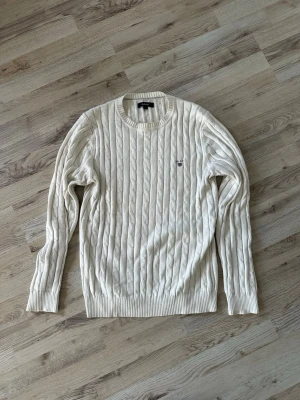 Vit kabelstickad tröja från Gant - Kabelstickad tröja i vitt från Gant med rund halsringning och ribbade muddar. Tröjan har ett broderat Gant-emblem på bröstet och är tillverkad i mjukt material. Perfekt för dig som gillar klassisk och stilren look.