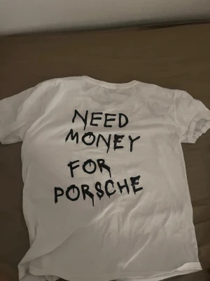 Vit t-shirt med tryck från BVALDI - Vit t-shirt från bvaldi med svart tryck på ryggen där det står 'NEED MONEY FOR PORSCHE' i handskriven stil. På bröstet finns en liten svart logga. T-shirten har rund hals och korta ärmar, perfekt för en chill och avslappnad look. Köpte den för 400 ny eller kanske till och med dyrare 