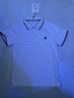 Vit pikétröja från Moncler - Vit pikétröja från Moncler med klassisk krage och knappar framtill. Tröjan har korta ärmar och diskreta ränder i rött och blått på krage och ärmslut. Moncler-logga broderad på bröstet. Perfekt för en stilren look.